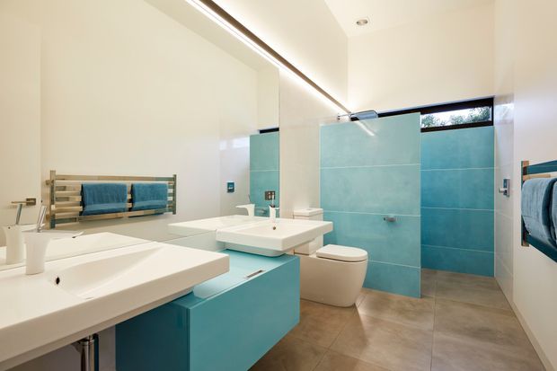 Bright contemporary bathroom with double white vessel basins, turquoise built-in vanity cabinetry, matching turquoise tiled shower walls, a freestanding toilet, large-format neutral floor tiles, wall-mounted towel rails, and a long illuminated mirror.