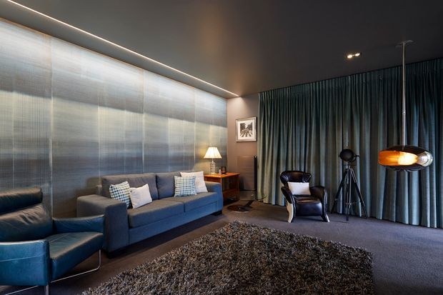 A moody, sophisticated living room or media lounge with deep blue upholstered seating, a textured dark rug, full-height blue-grey curtains, a metallic textured feature wall, and a dark ceiling with concealed linear lighting. A lounge chair, floor lamp, framed artwork, and sculptural floor lamp complete the refined interior.