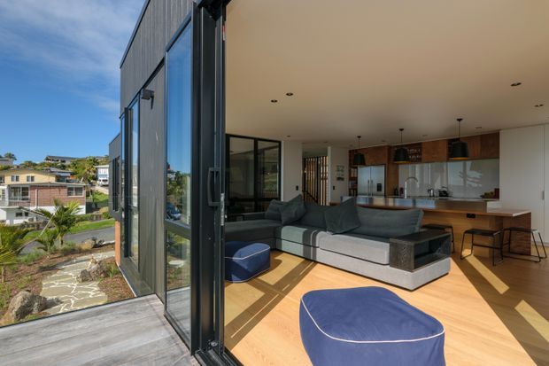 Bright contemporary open-plan living area viewed from a generous glazed exterior opening, with a grey sectional sofa and blue ottomans on a light timber floor. The kitchen beyond features warm timber cabinetry, a pale benchtop, pendant lights and integrated appliances. Full-height dark-framed glazing connects the interior to a small paved landscaped terrace and suburban surroundings.