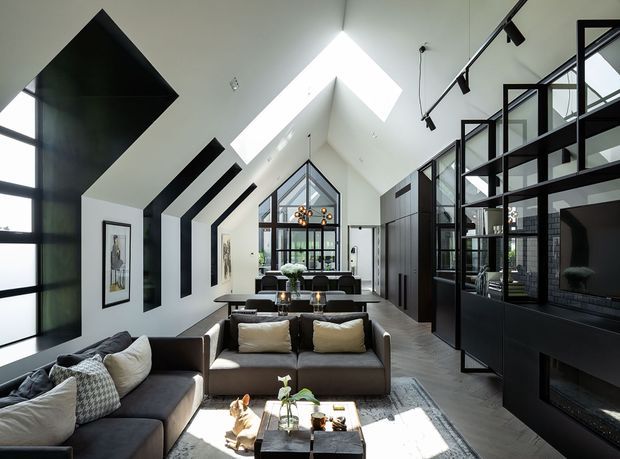 A sophisticated open-plan living room with a dramatic vaulted ceiling and large skylight. The space features dark built-in cabinetry and black-framed glazed partitions, a sectional sofa and central seating area, a feature fireplace or media wall, and a glazed gable at the far end. Strong black-and-white contrast is softened by warm timber-toned flooring and neutral upholstery.