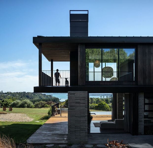 Modern two-storey dark-clad house on a landscaped sloping site, photographed from the exterior. The home features a strong horizontal flat-roofed form, deep covered balcony, extensive glazing, dark vertical timber cladding, a prominent chimney, and a timber deck extending toward a swimming pool and distant leafy landscape. The lower level opens to an outdoor living area, while a person and dog are visible on the upper balcony.