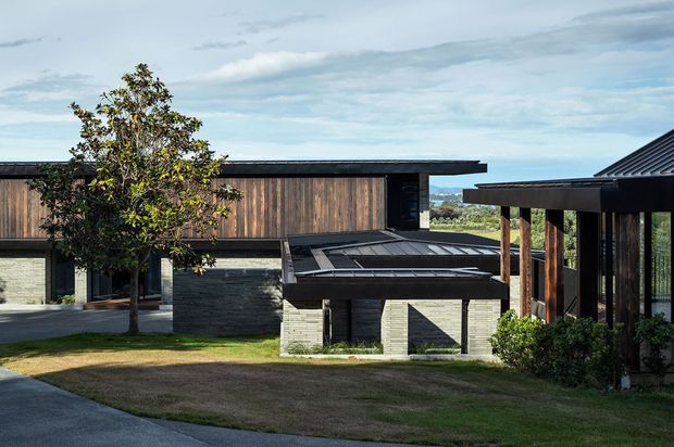 Exterior view of a contemporary low-rise residence composed of overlapping horizontal volumes. The building combines dark masonry, vertical timber cladding, broad flat roof planes, timber posts, and expansive glazing, with a landscaped lawn and a distant coastal or hilly outlook visible beyond.