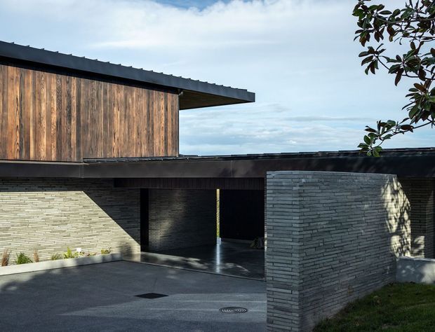 Exterior view of a contemporary low-rise residence with a strong horizontal composition. The building combines dark flat roof planes, weathered vertical timber cladding on the upper volume, and grey stacked masonry walls around a covered entry or carport. A paved forecourt and landscaped edges are visible, with clean minimal detailing and deep shadowed openings.