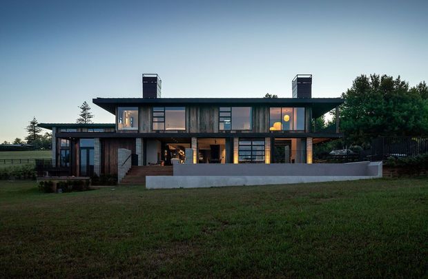 A contemporary two-storey residential house viewed from the front at dusk. The long, low-profile building has a broad flat roof with two chimney-like rooftop elements, extensive dark-framed glazing, timber-toned vertical cladding, dark structural framing, and a recessed upper balcony or terrace. A low pale concrete boundary wall and landscaped lawn sit in front, with mature vegetation around the property. Warm interior lighting contrasts with the cool blue evening sky.