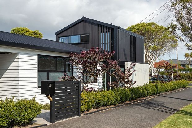 Street-facing exterior view of a contemporary two-storey residence combining a white horizontal weatherboard lower volume with a dark vertically clad gabled upper volume. The home has large rectangular windows, narrow vertical screening elements, a dark metal roof, a small slatted front gate and mailbox, and neatly clipped hedging along the paved frontage.