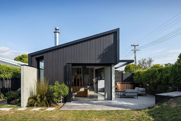 A contemporary single-storey dark-clad backyard extension or pavilion with a sharply sloping asymmetrical roofline. The black vertical cladding contrasts with pale rendered walls and extensive glazed doors opening onto a timber deck. The deck includes outdoor lounge seating and a compact timber hot tub or spa, with established hedging and residential fencing surrounding the landscaped yard. Utility poles and neighbouring suburban elements are visible beyond the property.