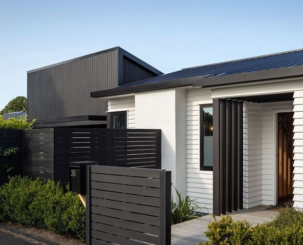 Exterior view of a contemporary single-storey home entrance, featuring white horizontal weatherboard cladding, a dark metal roof, black vertical and horizontal screening, and a recessed timber front door. Low clipped landscaping and a paved entry path frame the facade, with a taller dark-clad structure visible behind.