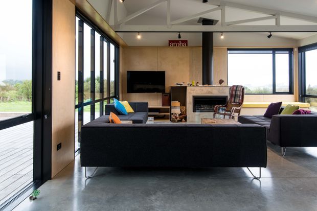 Open-plan contemporary living room with a large dark sectional sofa, additional lounge seating, a central stone fireplace, wall-mounted television, and extensive black-framed glazing opening to a deck and landscaped views. The room has a polished concrete floor, warm timber wall panelling, exposed white roof trusses, and a high, airy ceiling.
