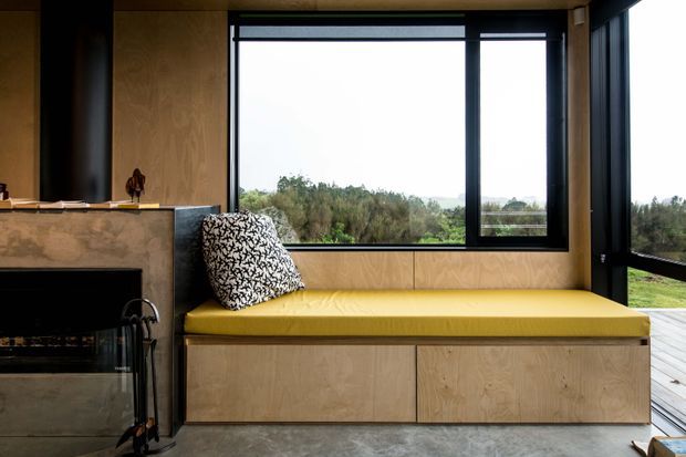 A compact contemporary sitting nook beside a black fireplace, featuring a built-in plywood window seat with mustard-yellow upholstered cushion and integrated storage drawers. Large black-framed windows overlook a wooded landscape, and a timber deck is visible outside.