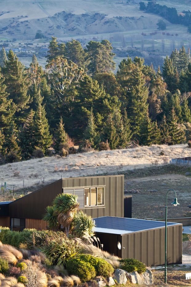 A modern dark-clad house set into a dry, hilly landscape, viewed from above through foreground trees and native plantings. The building has crisp rectilinear volumes, corrugated or ribbed metal wall cladding, a low-pitched metal roof, large windows, and a green street lamp beside the driveway. The surrounding setting includes coniferous trees, tussock-like grasses, rocky garden beds, and expansive rural hills.