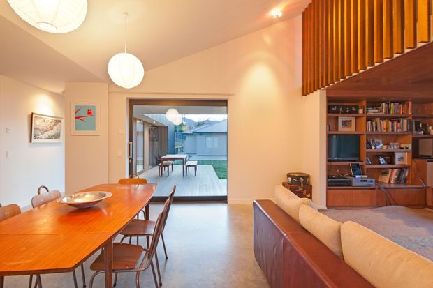 Warm, open-plan mid-century-inspired living and dining area with a polished timber dining table and chairs, sculptural pendant lights, a built-in timber entertainment and shelving unit, and a large upholstered sofa. Vertical timber slats define the upper section of the living area, while full-height glazing opens onto a paved courtyard and provides views toward neighbouring buildings.