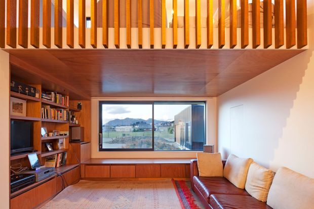 Warmly detailed living room with extensive built-in timber shelving and media joinery, a timber-lined ceiling, and a leather sofa facing the cabinetry. A full-height timber balustrade overlooks the room from an upper level, creating a mezzanine-like double-height feature. The large window frames a distant landscape and water view.