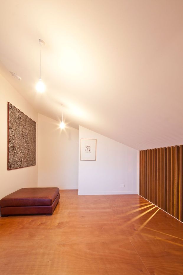Minimal, warm-toned interior with a polished timber floor, white walls and ceiling, a low brown leather ottoman, framed artwork, recessed and pendant lighting, and a vertical timber slat screen along the right side. The sloping ceiling and open floor area create a calm, contemporary lounge or living space.