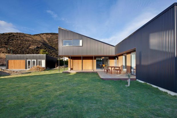 Exterior view of a contemporary single-storey house arranged around a lawn, with dark corrugated metal cladding, angular pitched roof forms, warm timber-lined walls, large glazed openings, and a covered outdoor terrace. A smaller matching outbuilding sits to the left, with dry rolling hills behind the property.