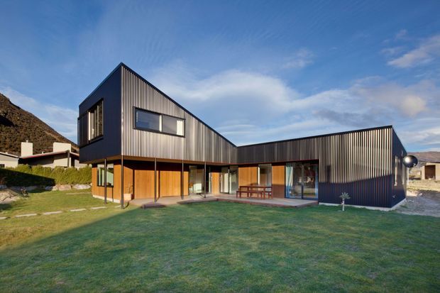 Exterior view of a contemporary multi-wing house with sharply pitched and low-slung roof forms, dark vertically ribbed metal cladding, extensive glazing, and warm timber-lined ground-floor walls. The home sits on a landscaped lawn with mountains visible in the background.