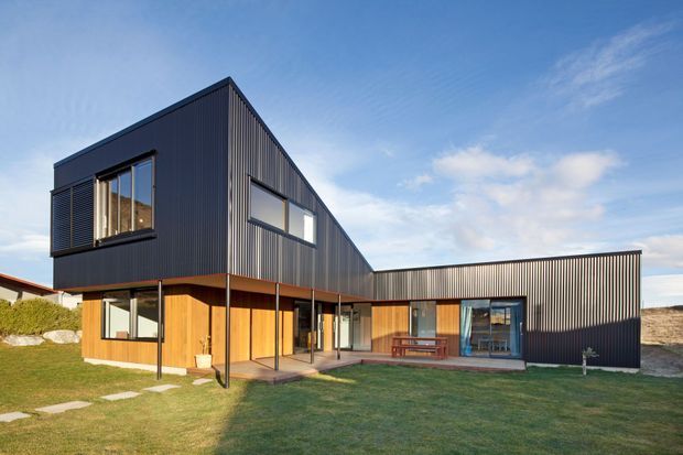 Exterior view of a contemporary multi-wing house with dark vertically ribbed metal cladding, warm timber wall panels, extensive glazing, and a low-pitched roof form. The home opens onto a broad lawn with a covered outdoor terrace and dining area.