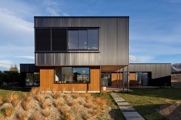 Exterior photograph of a contemporary two-storey residence composed of crisp rectangular volumes. The upper level is clad in dark vertically ribbed metal, with large glazed openings and projecting dark louvred screens. The lower level combines warm timber cladding, expansive windows and a covered entry walkway. A landscaped front garden with ornamental grasses and stepping-stone paving leads to the home, with open mountainous terrain visible in the distance.
