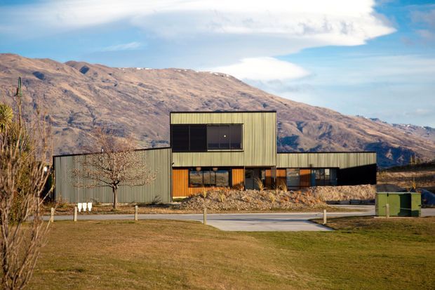 A low, angular contemporary building set against a rugged mountain landscape. The facade combines dark corrugated metal cladding with a raised central rectangular volume, broad horizontal glazing, and warm timber-toned entrance detailing. A paved approach and maintained lawn occupy the foreground.
