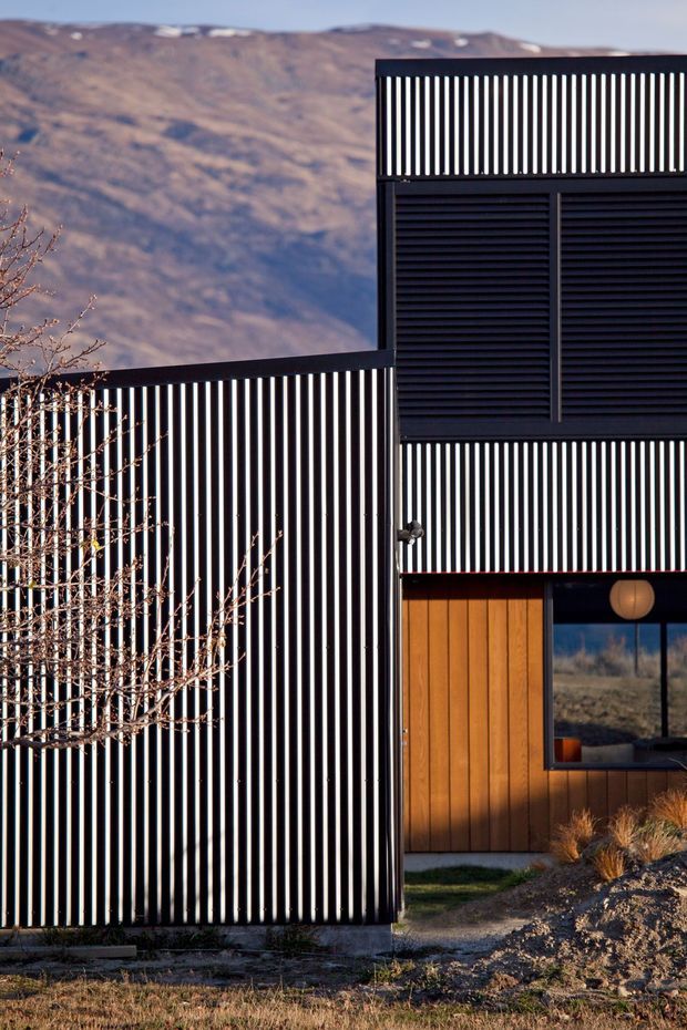 Exterior architectural photograph of a contemporary dark-clad building set against a rugged mountain landscape. The facade combines bold black vertical slatted screening, horizontal louvres, warm timber wall cladding, and a large glazed opening. A small tree and rough landscaped ground sit in the foreground.