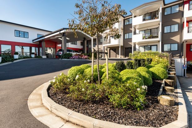 Exterior view of a contemporary multi-storey residential complex with a covered entry canopy, balconies, dark and light facade sections, and a landscaped central driveway island with clipped shrubs and young trees.