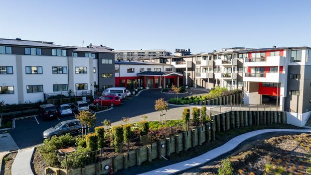 Aerial view of a contemporary multi-building residential apartment development arranged around a landscaped central driveway and entrance. The complex features white and grey façades, dark window frames, balconies, red accent panels and a sloping landscaped site with retaining walls and pedestrian paths.