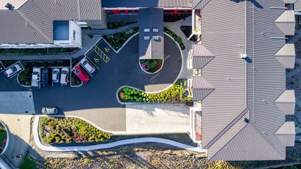 Aerial top-down view of a contemporary multi-building residential or commercial complex, showing grey metal pitched roofs, a central covered entry or service structure, curved asphalt driveways, landscaped garden beds, parking bays, and a small loop road.