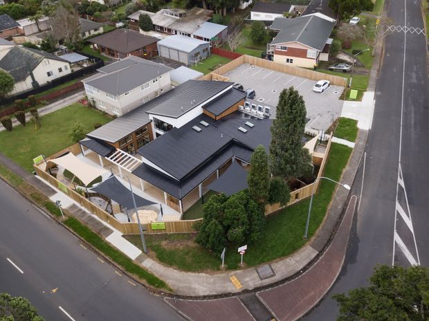 Aerial view of a contemporary single-storey residential house on a corner suburban site. The home has intersecting dark pitched roof forms, extensive glazing, timber-clad sections, paved outdoor areas, fenced landscaping, and a large adjoining driveway or parking area. Surrounding detached homes and streets are visible.