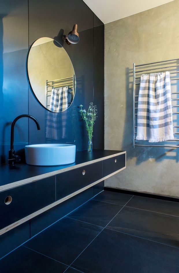 A contemporary bathroom featuring a floating dark vanity with integrated drawers, a white vessel basin, matte black curved tapware, a large round mirror, dark tiled flooring, and a chrome heated towel rail mounted on a textured warm-grey wall. Blue-and-white striped towels and a small vase of foliage add contrast.