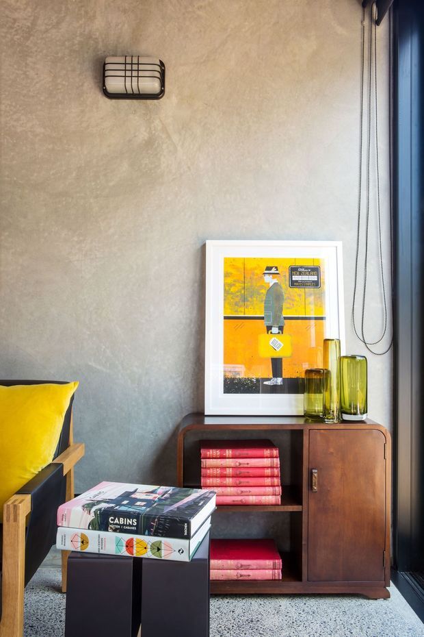A compact, stylish interior corner featuring a textured grey plaster wall, dark-framed full-height window, and a mid-century-inspired timber sideboard. The sideboard displays framed orange artwork, olive-green glass vessels, and stacked red books. In the foreground are a black square coffee table with design books and part of a timber-framed lounge chair with a bright yellow cushion. The space has a warm, eclectic, refined character with strong colour accents and natural light.