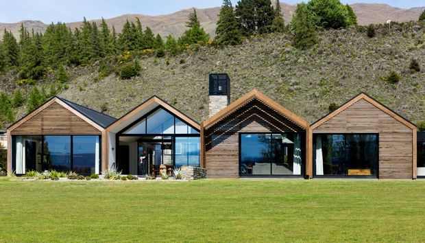 A single-storey contemporary mountain residence composed of several connected gabled volumes. The facade combines horizontal timber cladding, dark metal roofing, extensive floor-to-ceiling glazing, and a central stone-clad chimney. It sits at the edge of a broad lawn with rugged hills and evergreen vegetation behind it.