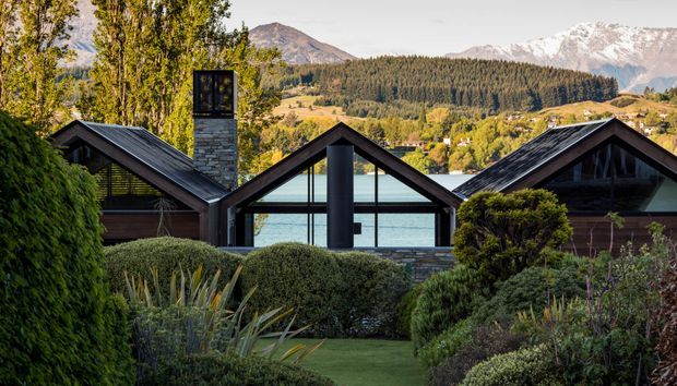 Exterior view of a low, dark-clad alpine residence composed of several steep gabled volumes, with a central glazed pavilion and prominent stone chimney. The home is set within a manicured garden, with a lake, forested hills, and snow-capped mountains visible beyond.