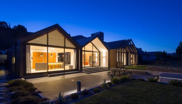 Twilight exterior view of a contemporary single-storey home composed of several connected gabled volumes. The façade features extensive floor-to-ceiling glazing, dark metal gable roofs, warm interior lighting, timber-framed glazing, and a broad paved terrace with landscaped planting and a small pool or water feature in the foreground.