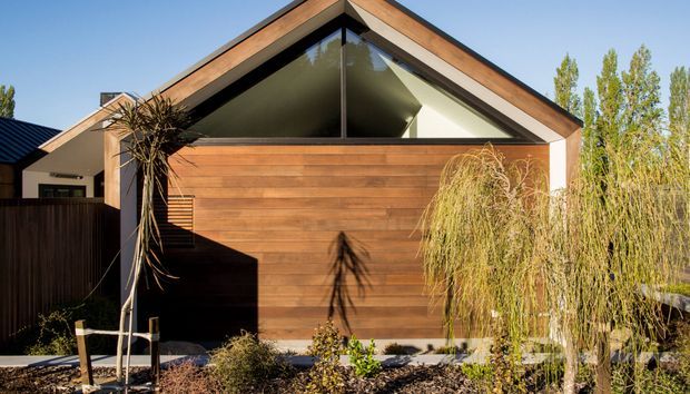 Front exterior view of a contemporary single-storey gable-roof house with warm horizontal timber cladding, a large triangular glazed gable, dark roof elements, and landscaped planting in the foreground.