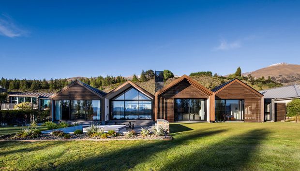 A row of low-rise contemporary residential pavilions with steep gabled roofs, dark timber cladding, extensive glazing, and landscaped lawns. The homes sit in a scenic mountainous setting, with a small landscaped pond or water feature in the foreground and a clear blue sky.