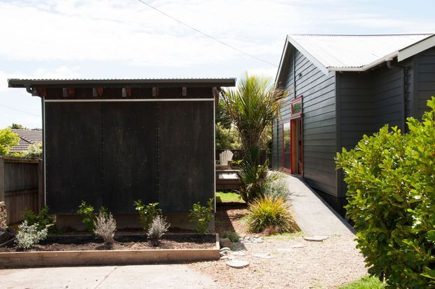 Exterior view of a compact dark-clad building with a corrugated metal pitched roof, red-framed glazing, and a narrow concrete path through a landscaped gravel garden. A separate dark timber outbuilding or screening structure sits in the foreground, with established shrubs and spiky ornamental planting around the property.