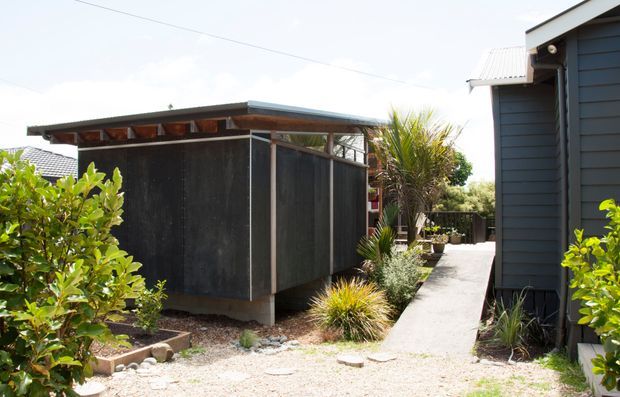 Exterior view of a compact contemporary residential property, showing a dark timber-clad building on the right and a separate charcoal-clad flat-roofed structure with clerestory-style openings. A concrete pedestrian path runs between the structures, bordered by low-maintenance ornamental planting and gravel landscaping.