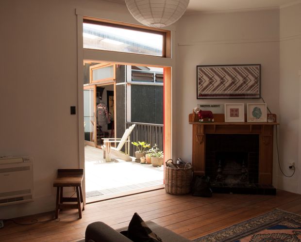 Warm, character-filled living room with timber floorboards, a traditional carved timber fireplace mantel, framed artwork, and a large glazed opening leading to a bright courtyard or deck. A woven pendant light, patterned rug, potted plants, and vintage furnishings create a relaxed eclectic atmosphere. The opening includes a high-level transom window and timber-framed glazing.