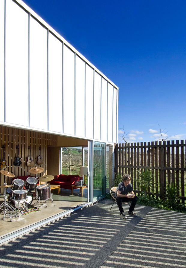 Exterior view of a contemporary multi-level residential building with a crisp white vertically panelled upper facade, extensive floor-to-ceiling glazing, and a partially enclosed ground-level outdoor entertaining area. The lower space contains a drum kit, guitars, and seating, while a dark timber slat fence and paved terrace occupy the foreground. Strong sunlight creates pronounced striped shadows across the terrace.