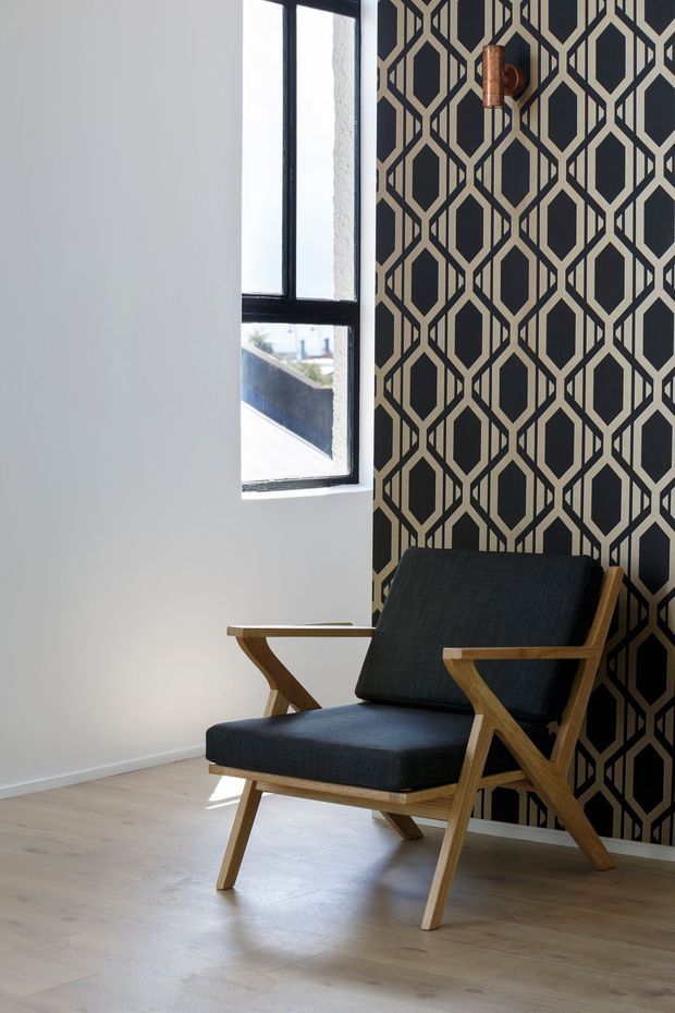 A bright interior corner featuring a mid-century-style wooden lounge chair with dark charcoal upholstery, positioned against a bold black-and-beige geometric feature wall. A black-framed window provides natural light, and pale timber flooring and white walls create contrast.