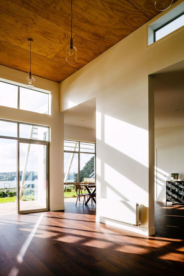 Bright, double-height modern interior with expansive glazing, a warm plywood-lined ceiling, dark timber flooring, and white plastered walls. Strong sunlight casts geometric shadows across the open-plan space. A small dining area is visible near the windows, with an exterior landscape and water view beyond the glazing.