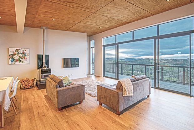 A bright, spacious living room with large floor-to-ceiling glazed doors and windows opening to a balcony with expansive landscape views. The room features warm timber flooring and a matching timber-panelled ceiling, neutral walls, two upholstered armchairs, a small fireplace or wood-burning stove, and contemporary artwork. A dining area is partially visible at the left edge.