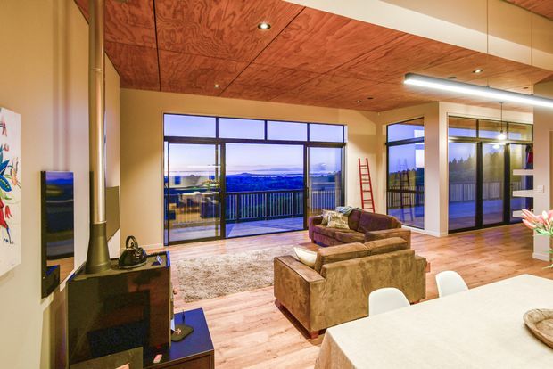 Warm contemporary open-plan living and dining area with expansive floor-to-ceiling glazing opening to a balcony and distant landscape views. The room features a timber-lined ceiling, timber flooring, a dark freestanding wood-burning stove, comfortable upholstered sofas, and a large dining table. Recessed and linear lighting create an inviting evening atmosphere.