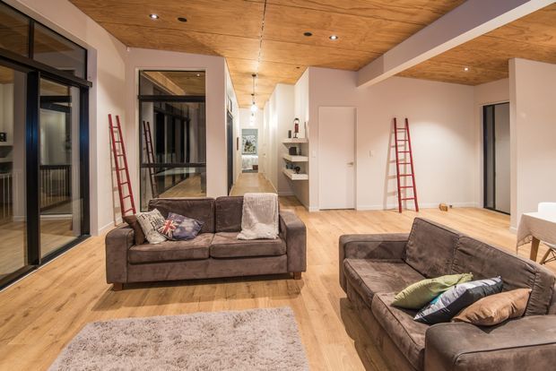 Spacious open-plan living area with two brown sofas, a pale textured rug, timber flooring, and a warm timber-lined ceiling. Full-height black-framed glazing and internal glass partitions line the left side, while a long hallway with built-in shelving and white doors extends into the background. Red ladders are visible in the room, suggesting the space may be under final fit-out or renovation.