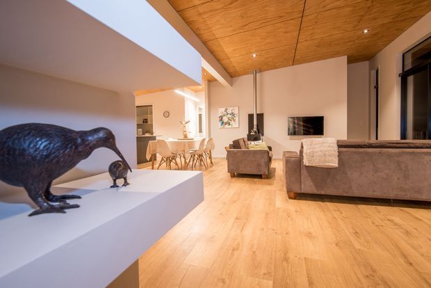 Open-plan contemporary living and dining area with warm timber flooring and a striking timber-lined ceiling. A large sectional sofa faces a wall-mounted television, while a dining table and chairs sit beyond near built-in shelving. White sculptural display plinths with bird forms occupy the foreground, and expansive dark-framed glazing is visible to the right.