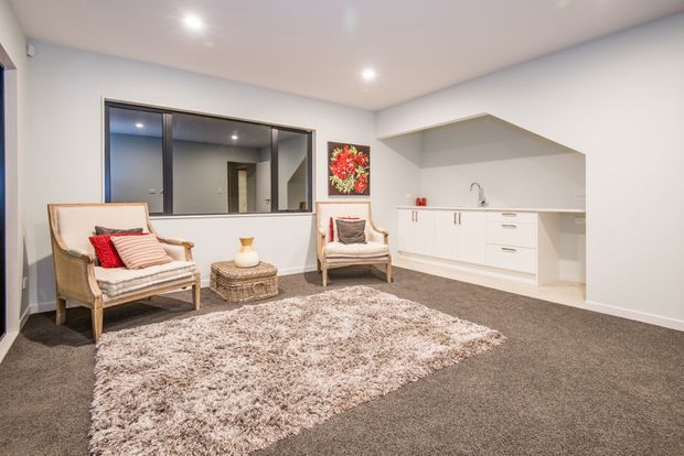 Bright, contemporary open-plan living area with two upholstered armchairs, a woven coffee table, a large shag rug, and a compact white kitchenette along the back wall. A dark-framed internal window, recessed ceiling lights, and neutral finishes create a clean, comfortable space.