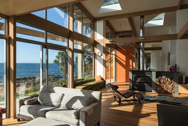 A bright, double-height coastal living room with expansive floor-to-ceiling glazing overlooking the ocean and rocky shoreline. The space features exposed timber beams, a warm timber floor, skylights, a grey sofa, green built-in window seating, a modern fireplace with stacked firewood, and strong natural sunlight.