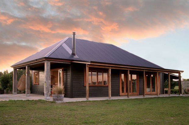Single-storey dark timber-clad house with a broad hipped corrugated metal roof, timber verandah framing, exposed posts, and extensive glazed doors and windows. The home sits in a simple grassy setting beneath a dramatic sunset sky.