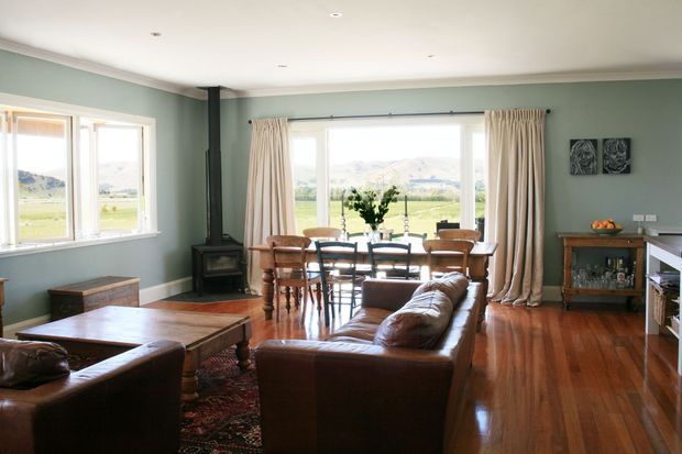 A bright, traditional open-plan living and dining room with muted sage-green walls, white trim, polished timber flooring, leather sofas, a timber dining table and chairs, and a freestanding wood-burning stove. Large windows and glazed doors provide expansive views of a rural valley and mountains, while full-length neutral curtains frame the central opening. A timber drinks cabinet and framed artwork complete the warm, classic interior.