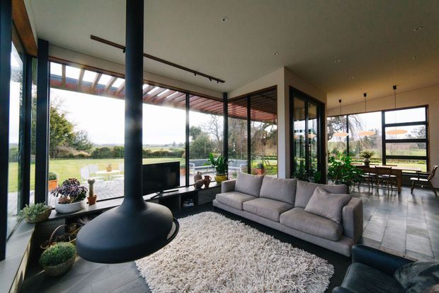 Contemporary open-plan living room with floor-to-ceiling glazing overlooking a landscaped rural setting. The room features a suspended black freestanding fireplace, large grey sectional sofa, shag rug, dark tiled flooring, exposed timber pergola-like glazing structure, and an adjacent dining area with extensive windows.