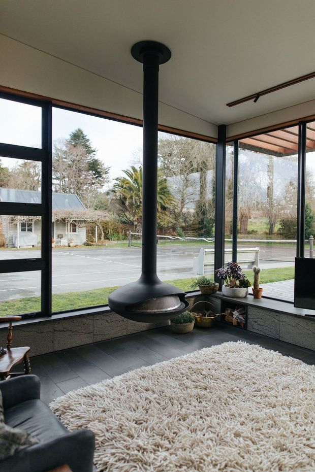 A mid-century-inspired living room with expansive black-framed floor-to-ceiling glazing, a suspended black freestanding fireplace, dark tiled flooring, a low textured masonry wall, and a large cream shag rug. Indoor plants and a black leather lounge chair add warmth, while a covered outdoor area and landscaped setting are visible through the windows.